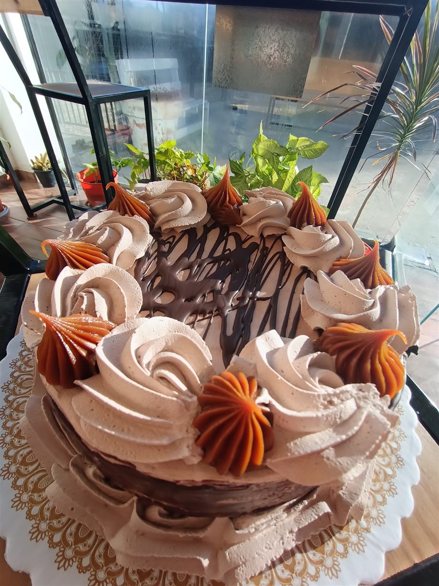 Torta de crema cacao con una corona de rosetas de crema alternadas con flores de dulce de leche, y un hilo de ganache dibujado sobre el centro, servida sobre un blondín dorado.