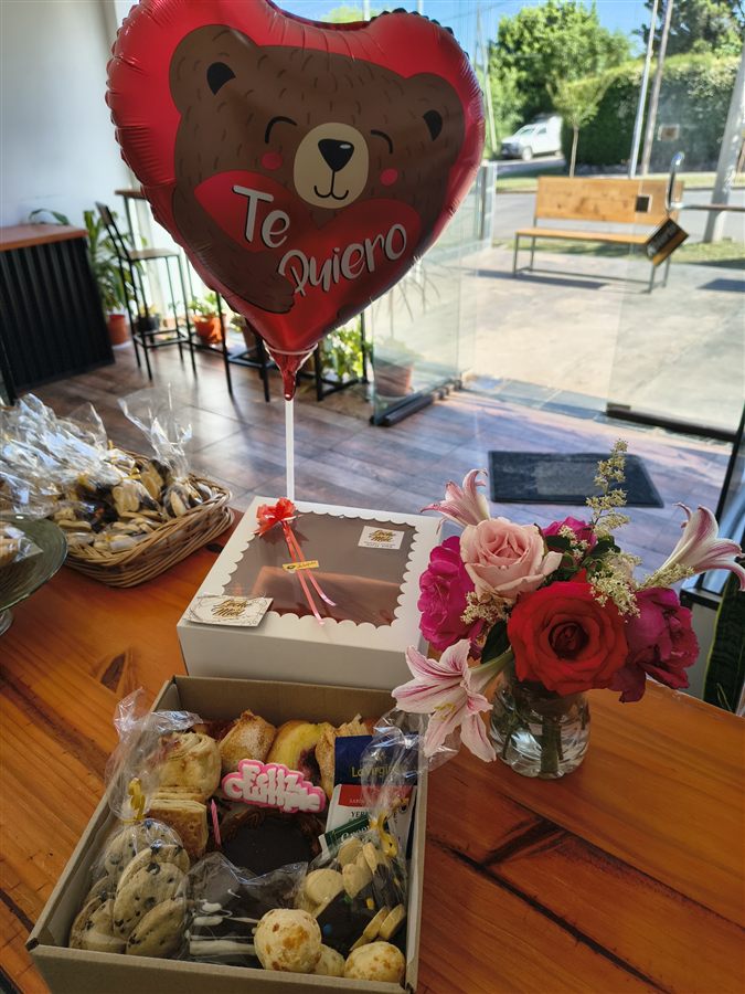 Caja de regalo con el sticker de Leche & Miel sobre la tapa, flores frescas al lado y una canasta con facturas al fondo.