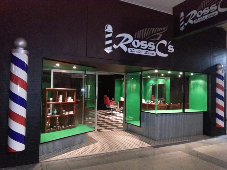 Ross C's illuminated shopfront at night with its two barber poles and green interior glow