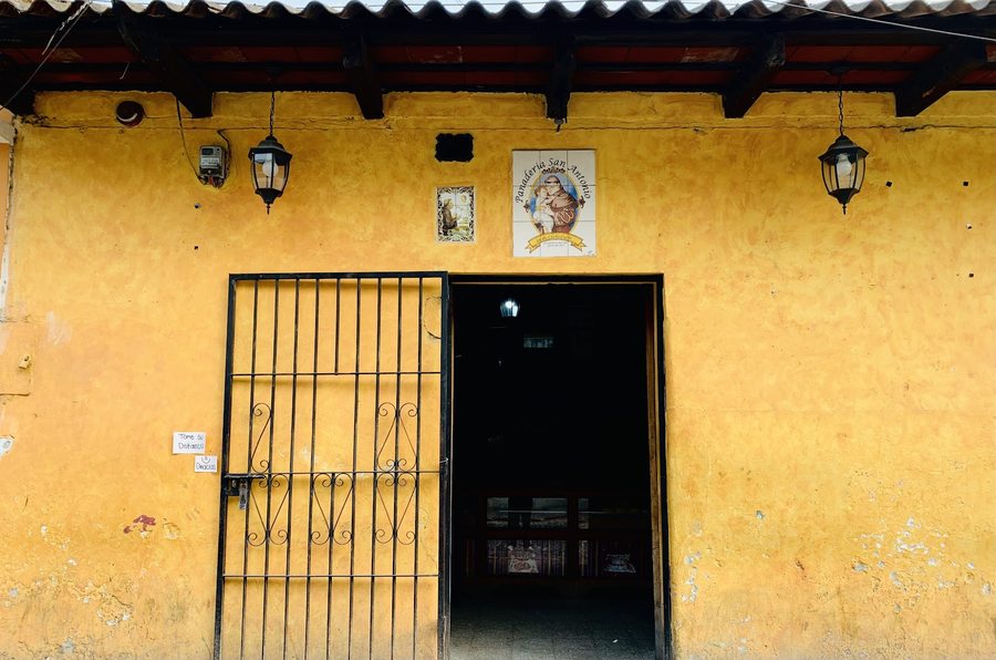 Fachada colonial ocre de la panadería con rótulo de azulejos y portón de hierro, en la 5a Calle Poniente de Antigua Guatemala