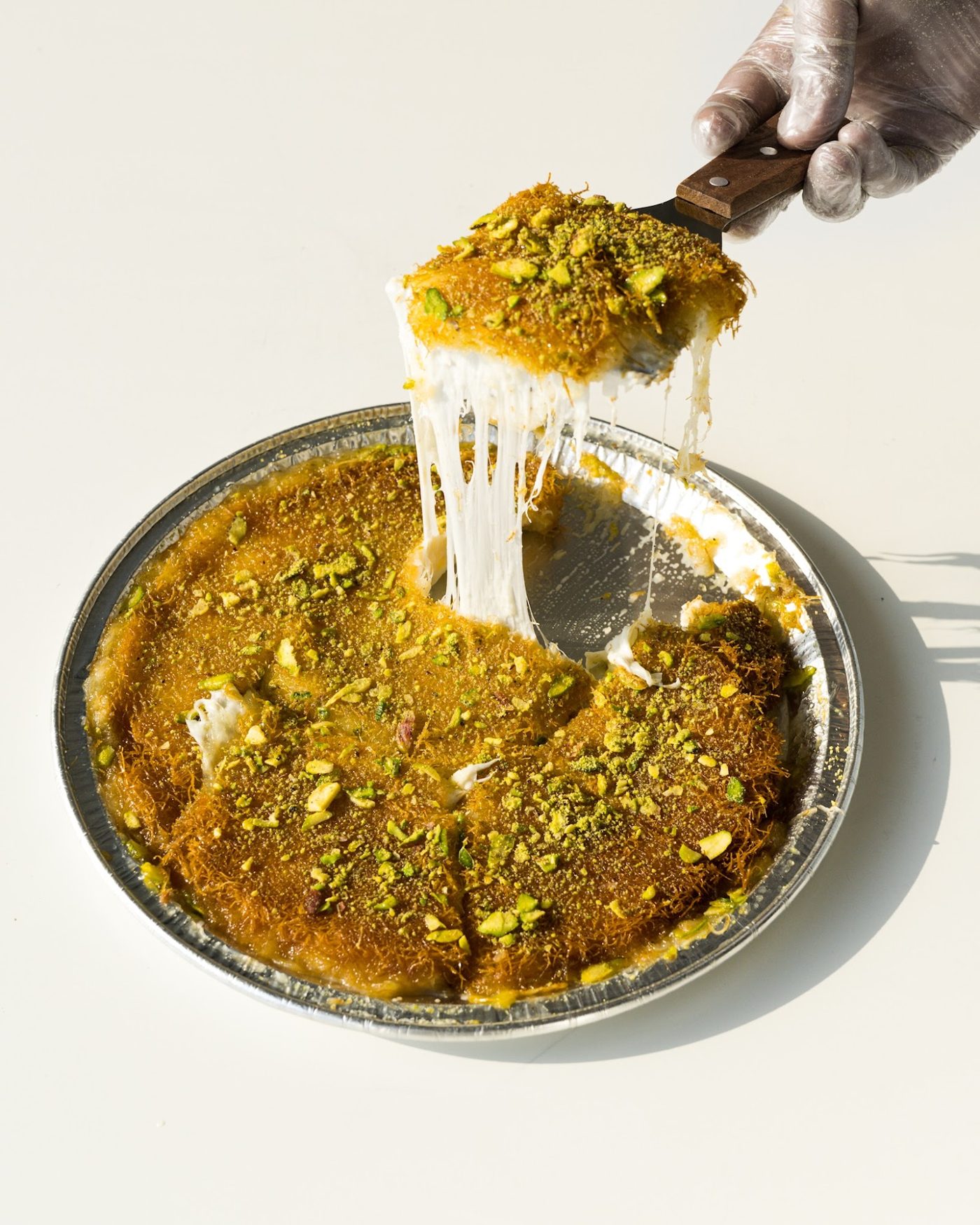 Tray of golden kunafa topped with pistachio, a slice lifted with a cheese pull