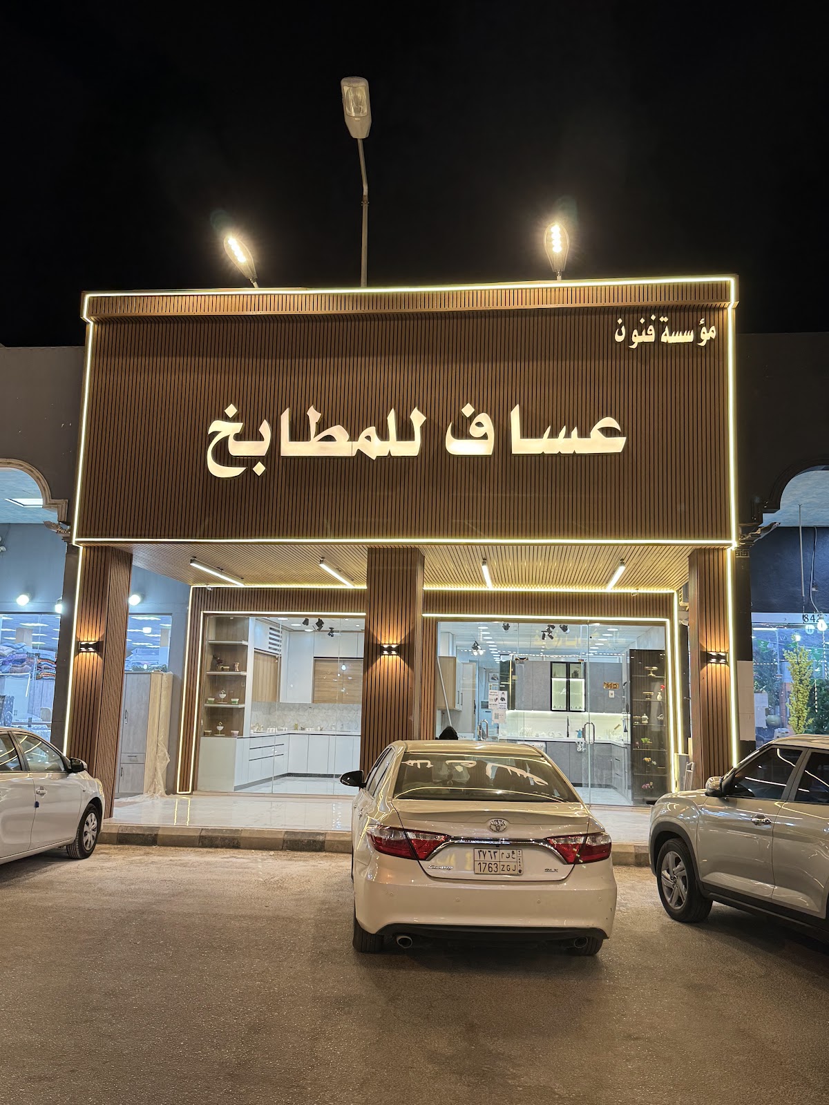 واجهة معرض عساف للمطابخ ليلًا بألواح خشبية طولية وإطار إضاءة دافئ، ومطابخ العرض خلف الزجاج