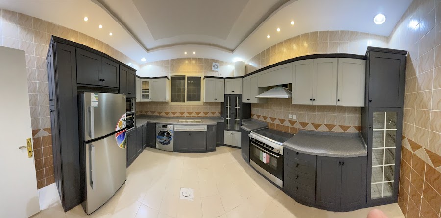 مطبخ واسع على شكل U بدرف شيكر فحمية سفلية ودرف علوية فاتحة، وخزانة عرض زجاجية بتقسيمات بيضاء وسطح رمادي