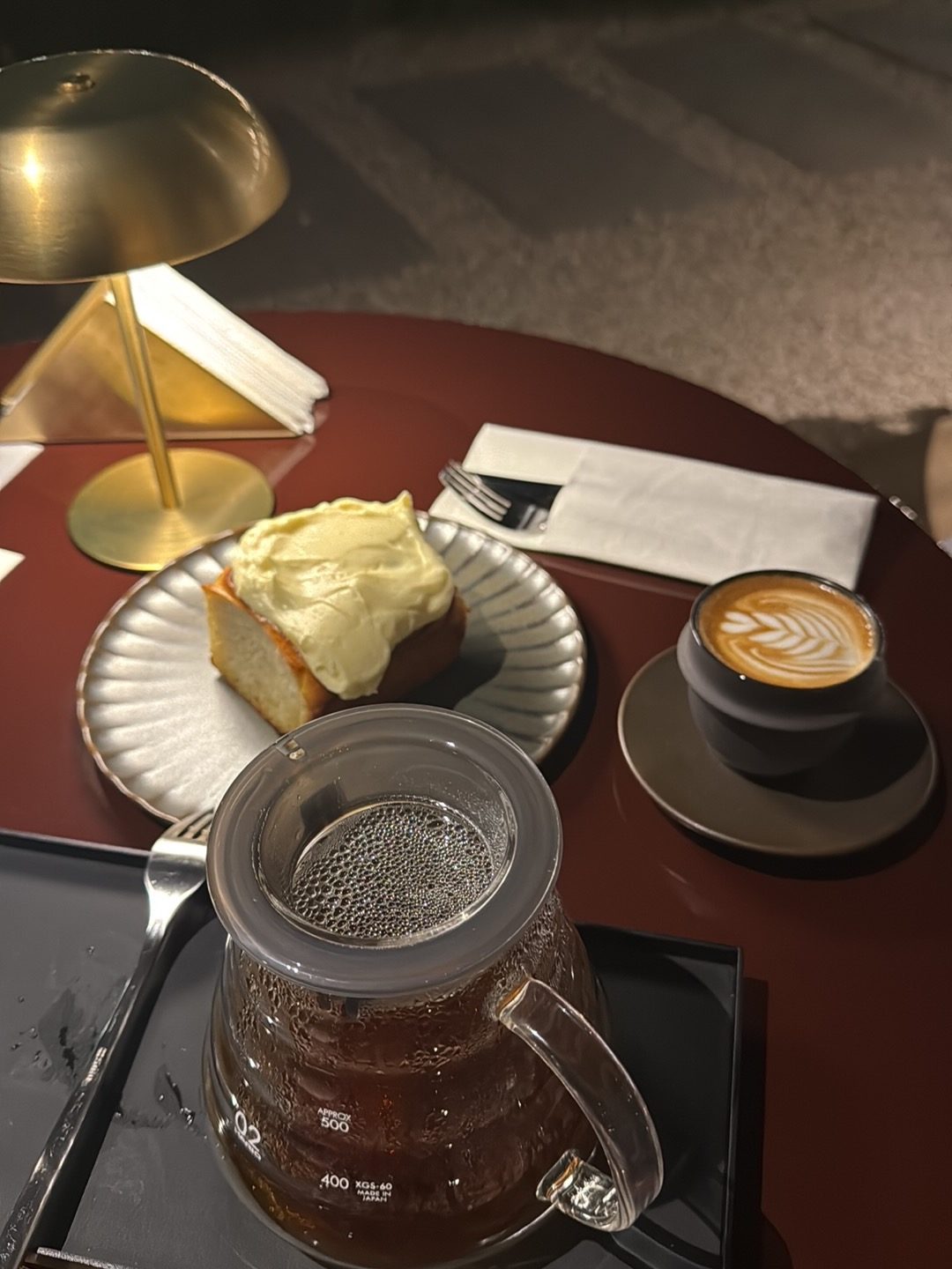 A rosetta-art flat white, a cream-topped cake slice, and a glass pour-over carafe on an oxblood table under a brass lamp