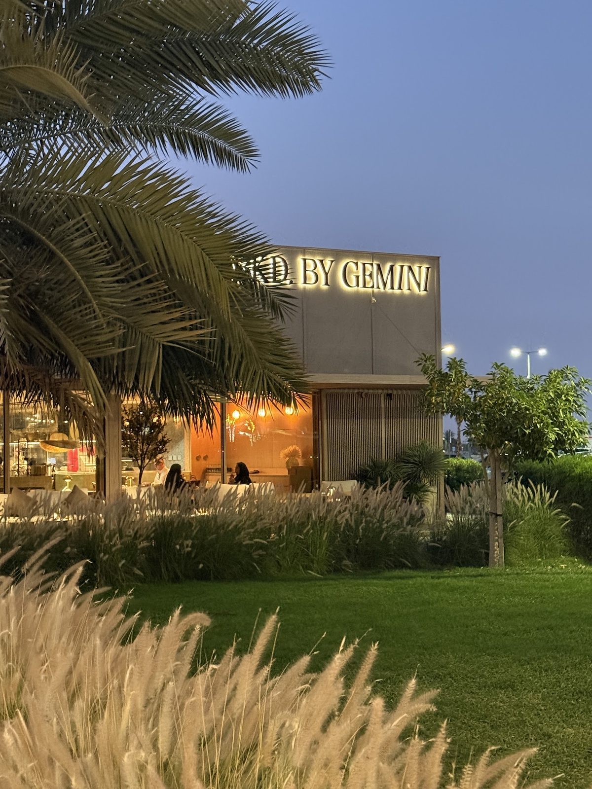 The lit BKD. BY GEMINI façade signage at dusk amid palms and ornamental grasses