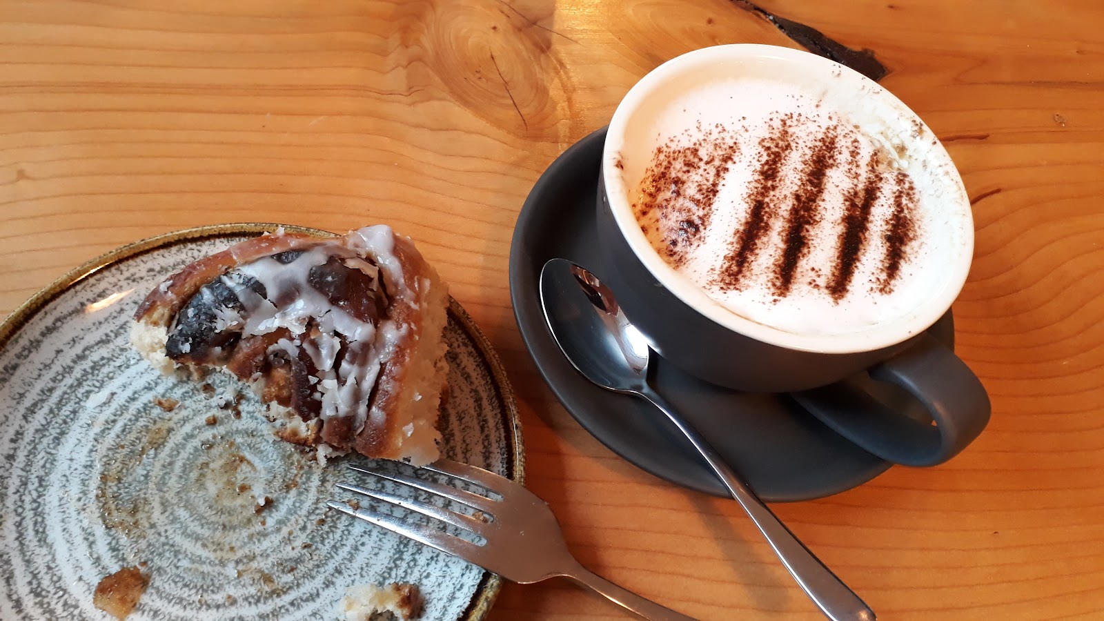 An iced cinnamon bun on a ceramic plate beside a cappuccino