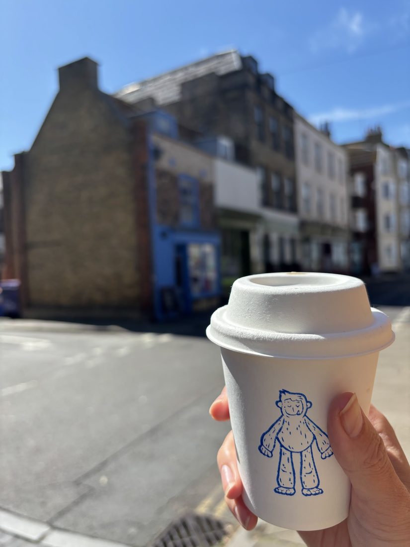 A GIANT COFFEE takeaway cup, printed with the shop's hand-drawn blue giant, held up on a Margate Old Town street.