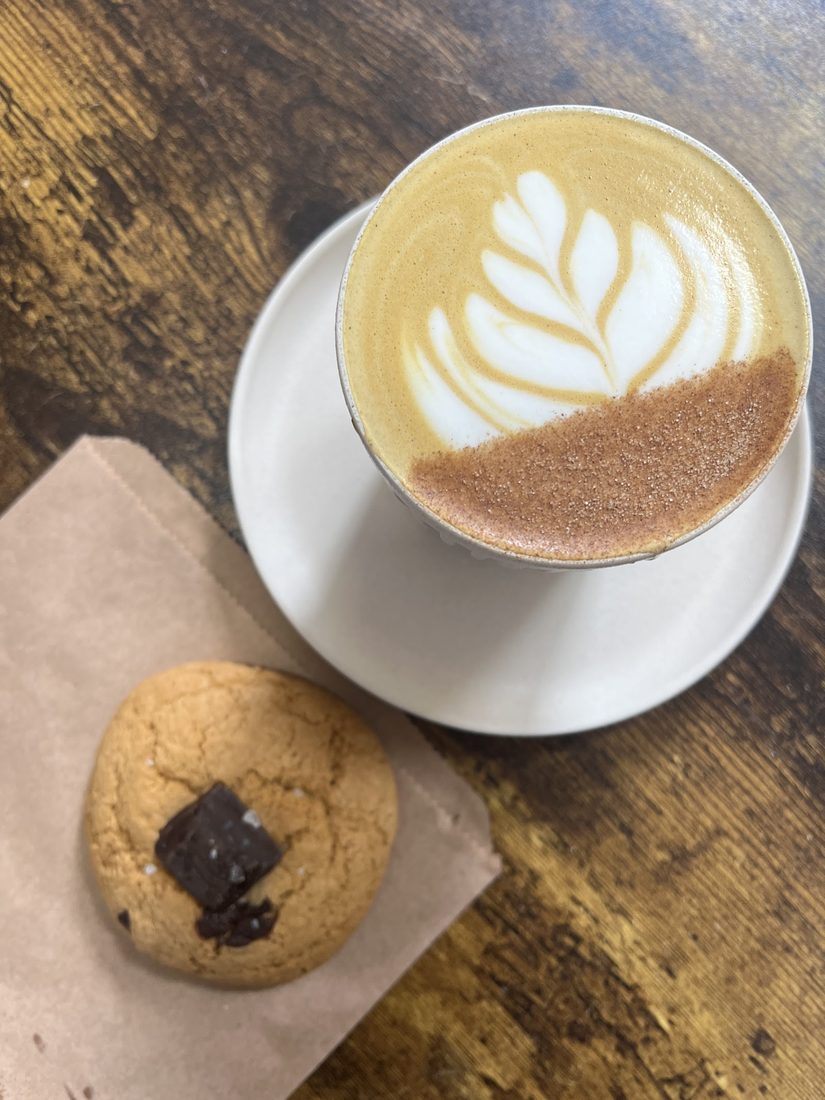 A flat white with fern latte art beside a chocolate-chunk cookie on a wooden table at GIANT COFFEE.