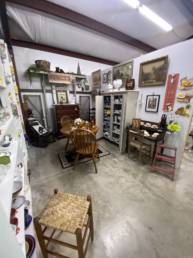 Inside a vendor booth — an oak table and chairs, a white milk-glass cabinet and framed landscape art.